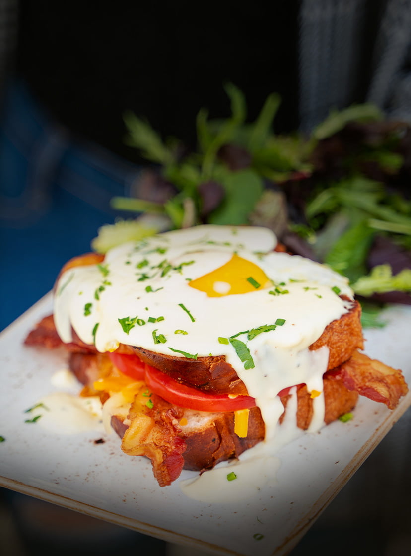 Modern Croque Madame, Our take on a French Classic. Challah (hall-a) bread griddled with melted Cheddar and Monterey Jack cheese, fresh tomato, and crispy bacon topped with a cage-free sunny side-up egg, Parmesan cream sauce and fresh herbs. Served with lemon-dressed organic mixed greens.